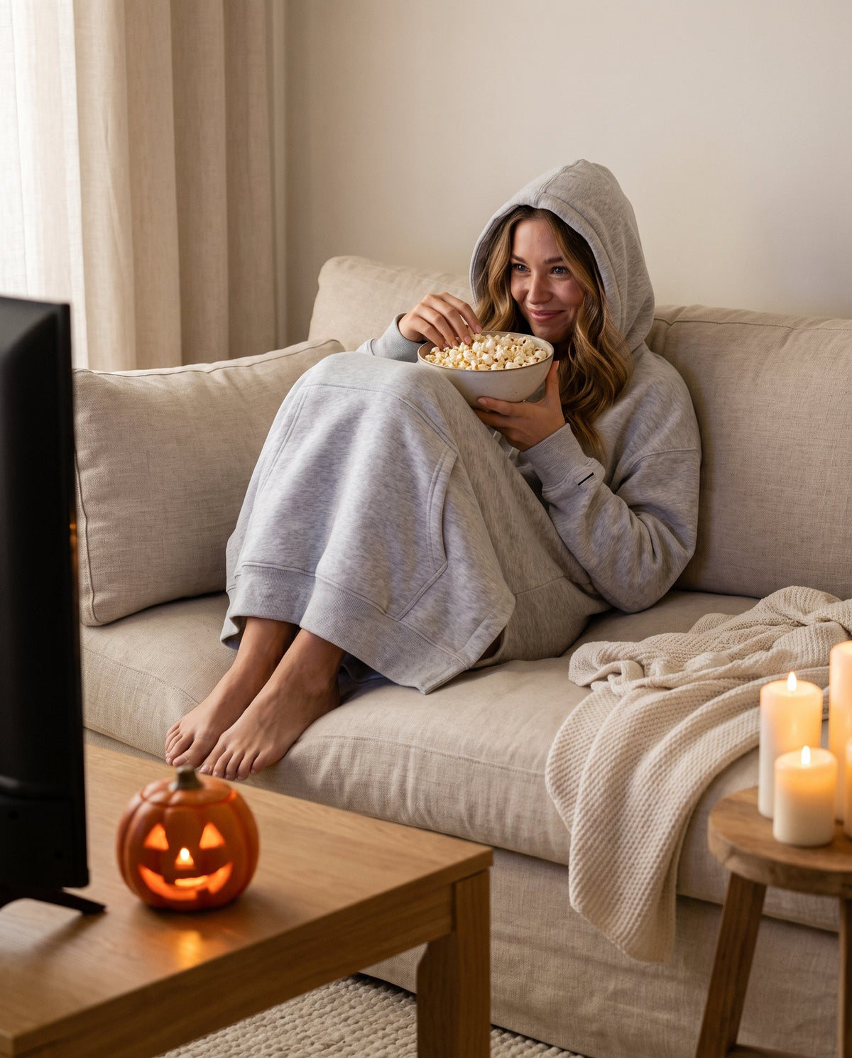 Woman curled up on the sofa for a Halloween movie night in the heather grey Junemae hoodie dress, hood up, with popcorn