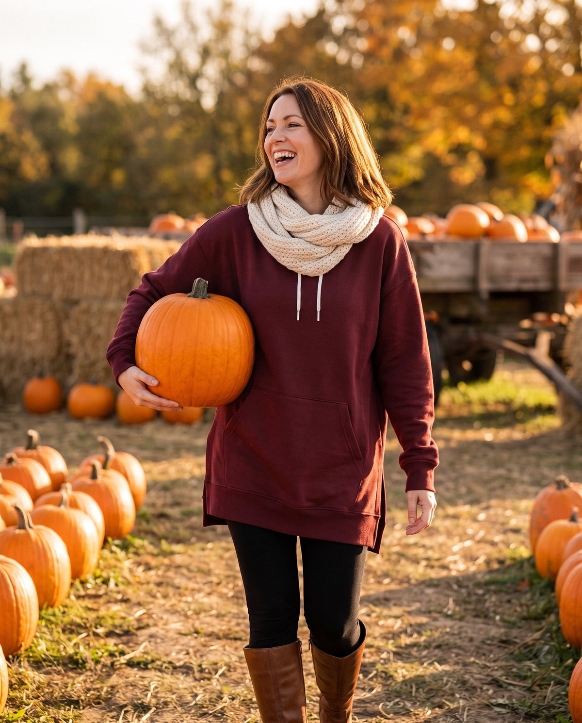 Woman carrying a pumpkin at a pumpkin patch in the burgundy Junemae hoodie dress, leggings and knee-high boots